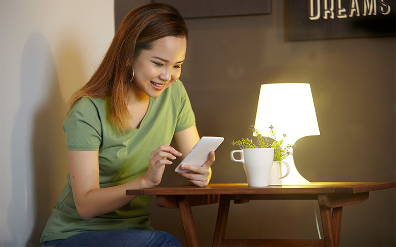 Young woman smiling while using smartphone at a cozy table with a cup and lamp.