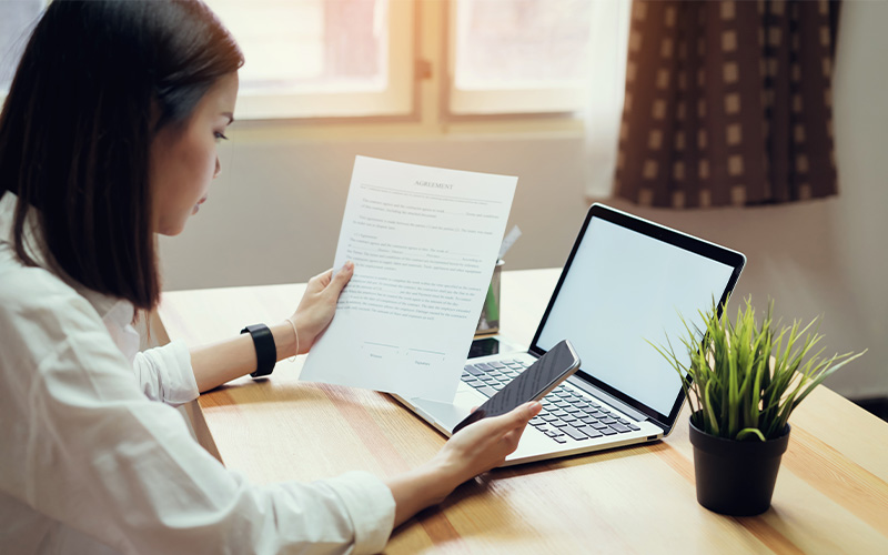 Woman reading a document while holding a phone, seated at a desk with a laptop and potted plant.