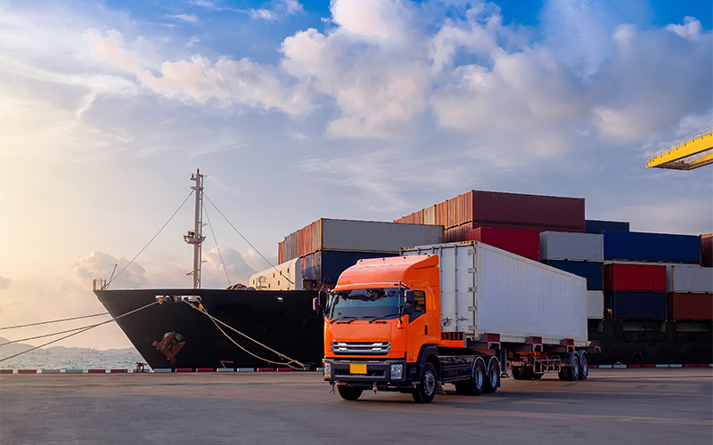 Orange cargo truck parked at a shipping port with stacked containers and a cargo ship in the background