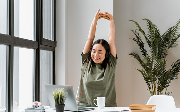 A woman wearing headphones stretches her arms above her head at a desk with a laptop.
