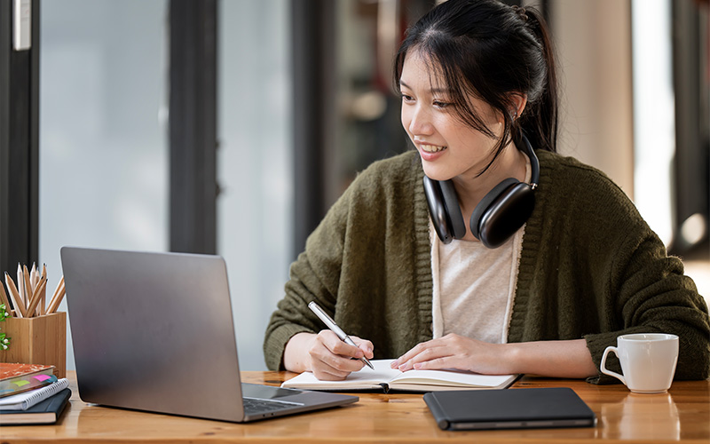 girl wearing headphones writing on her notebook while looking at the laptop